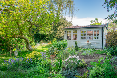 Langham Studio Home Study Grey timber garden office surrounded by mature trees and colourful planting, with a small table and chairs outside.